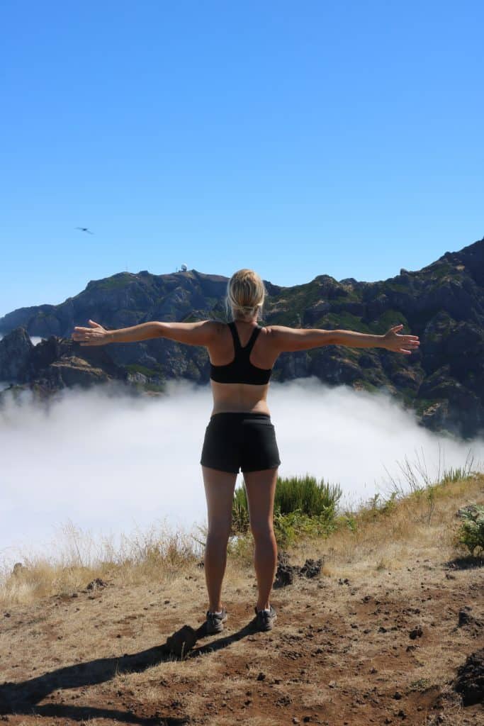 Girl in sport's top and short spreading her arms on top of a mountain in front of clouds