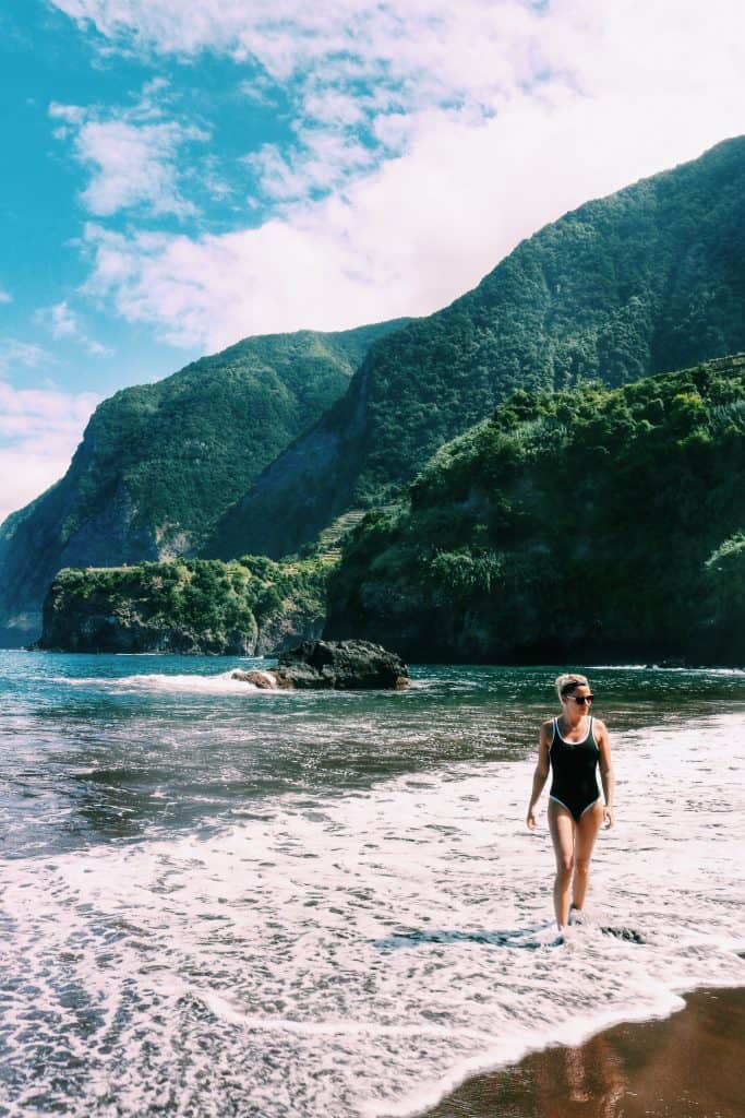 Girl walking in the ocean with a black swimming suit and a green mountain behind her