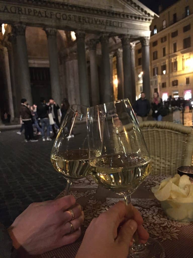 Two hands holding two glasses of white wine in front of pantheon