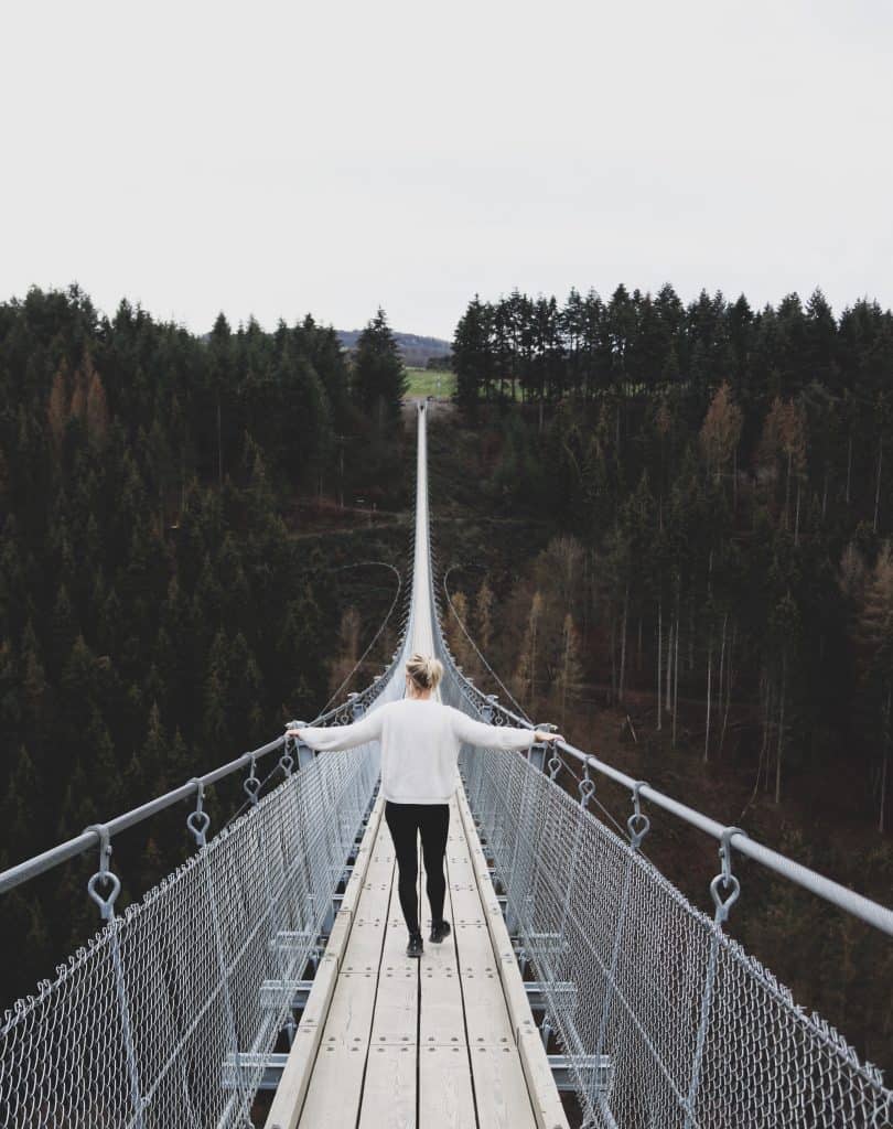 Girl with white sweater crossing a long bridge surrounded with pine trees