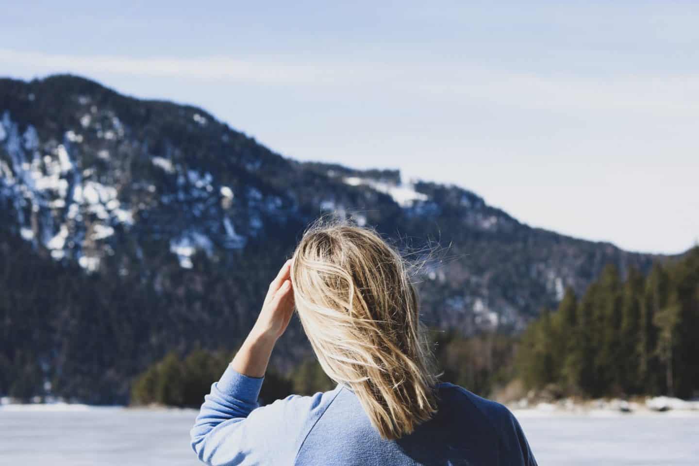 Girl with blonde hair standing in front of a snowy mountain with pine trees