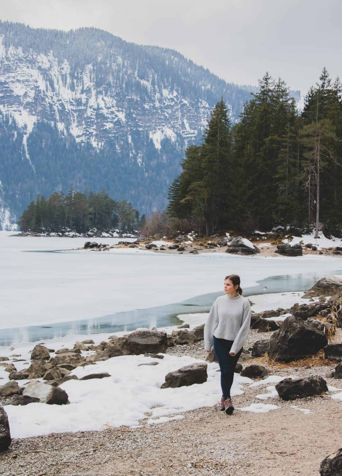 Girl walking next to a frozen lake with a mountain and pine trees behind